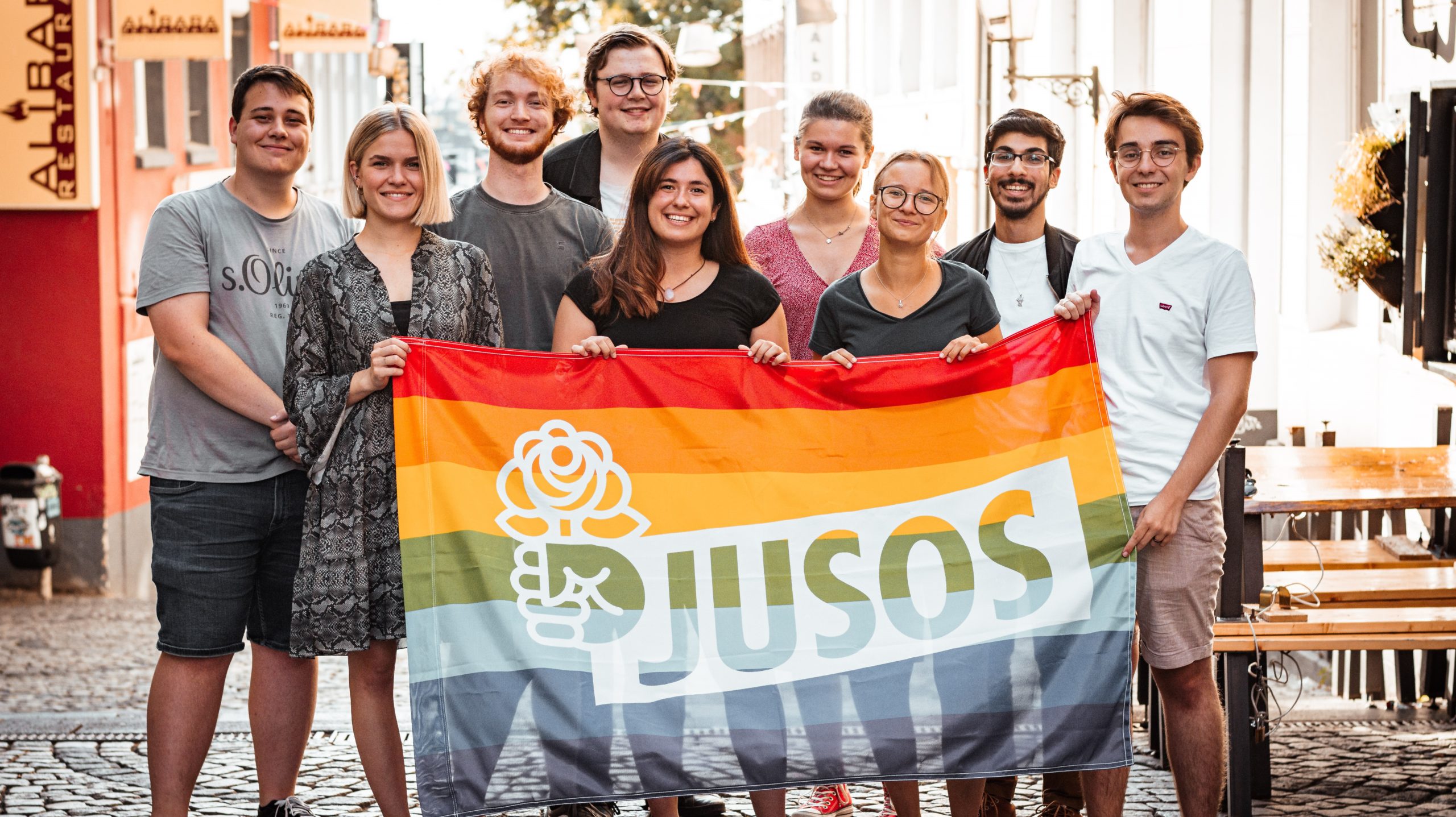 Bild, mit allen Vorstandsmitgliedern, auf dem sie eine Pride-Flagge halten. Auf der Pride-Flagge ist das Zeichen der Jusos abgedruckt.